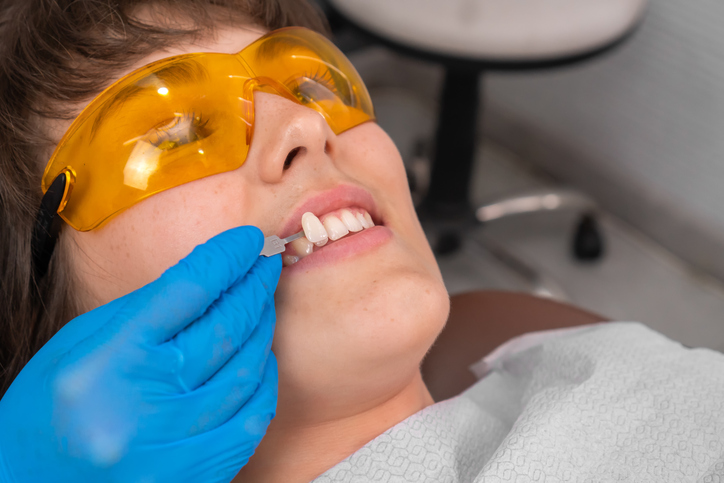 Girl patient and dentures teeth shade selection. Dentist selecting tooth color from special scale. Young woman sitting in dental operation chair. Prosthodontics. Bleaching .